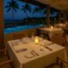 Candlelit dinner table set by the pool with palm trees and ocean view at a tropical resort restaurant at night.