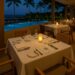 Candlelit dinner table set by the pool with palm trees and ocean view at a tropical resort restaurant at night.