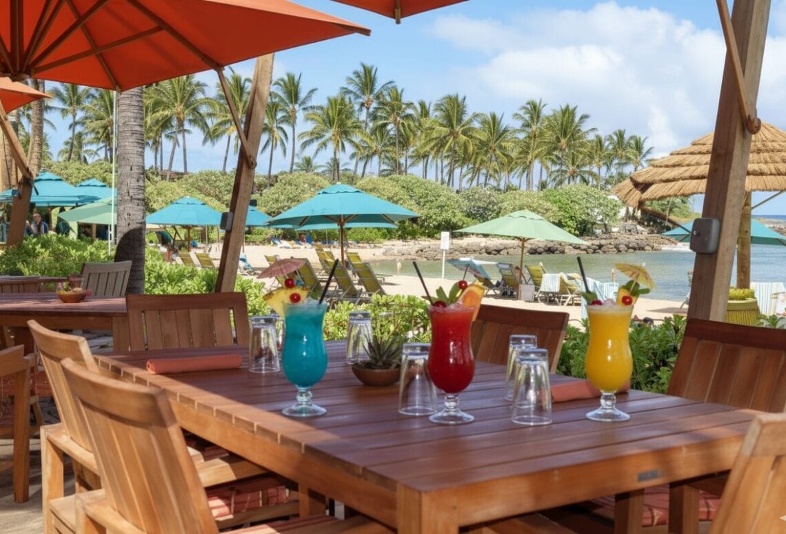 Wooden dining table with Hawaiian cocktails and beach chairs under umbrellas at The Beach House Restaurant, Turtle Bay, Oahu.