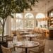 An elegant, light-filled dining room at Fig Restaurant in Santa Monica with high ceilings, hanging plants, communal tables, and a view of the garden patio.