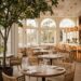 An elegant, light-filled dining room at Fig Restaurant in Santa Monica with high ceilings, hanging plants, communal tables, and a view of the garden patio.