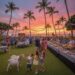 Three children interact with two small goats on a grassy lawn during a 4th of July celebration at a tropical resort at sunset. In the background, a crowd of people enjoys a buffet and live music under palm trees decorated with patriotic streamers and tiki torches.