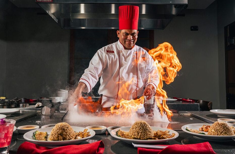 A smiling teppanyaki chef in a white uniform and tall red hat creates a large fire flare on a flat-top grill, with plates of fried rice and vegetables lined up in the foreground.