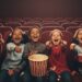 A group of six diverse children, four boys and two girls, sitting in the front row of a dark movie theater. They are all laughing and pointing excitedly. One child in the middle holds a large bucket of popcorn.