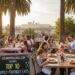 A vibrant, high-angle shot of multiple families with young children and teenagers enjoying meals at an outdoor restaurant patio in Los Angeles. The tables are set with checkered red and white tablecloths. In the background, the downtown Los Angeles skyline is visible at sunset, framed by tall palm trees. A chalkboard sign in the foreground reads "CulinaryPassages.com Presents: Top 10 Family-Friendly Eats - Los Angeles."