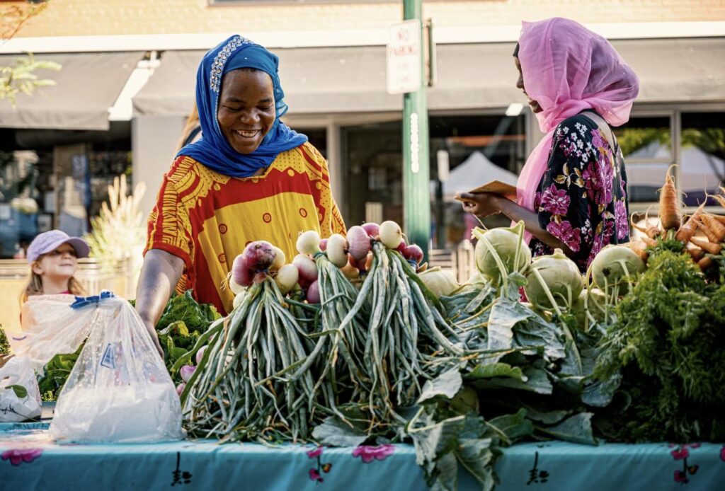 Two vendors at an outdoor farmers market stand piled with fresh turnips, kohlrabi, carrots, and greens, with a young girl looking on.