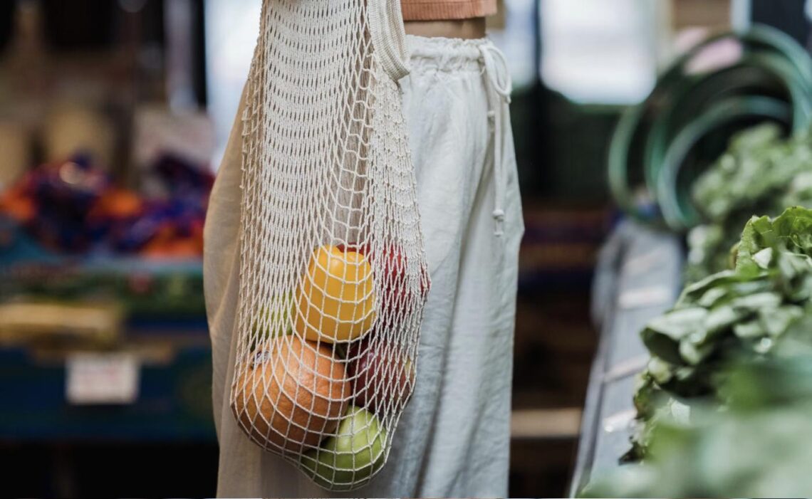A woman carries a mesh tote bag filled with fresh fruit at an indoor farmers market.