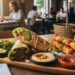 A wooden platter featuring two falafel wraps, grilled chicken skewers with bell peppers, a side salad with lemon wedges, creamy hummus topped with chickpeas, and a red dipping sauce, set on a restaurant table with a blurred interior background.