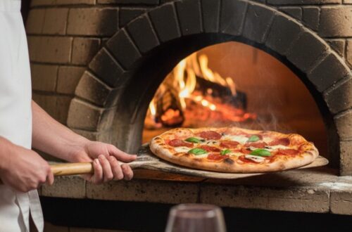 A chef in a white apron uses a wooden pizza peel to remove a fresh pepperoni and basil pizza from a traditional brick wood-fired oven with a visible roaring flame inside.