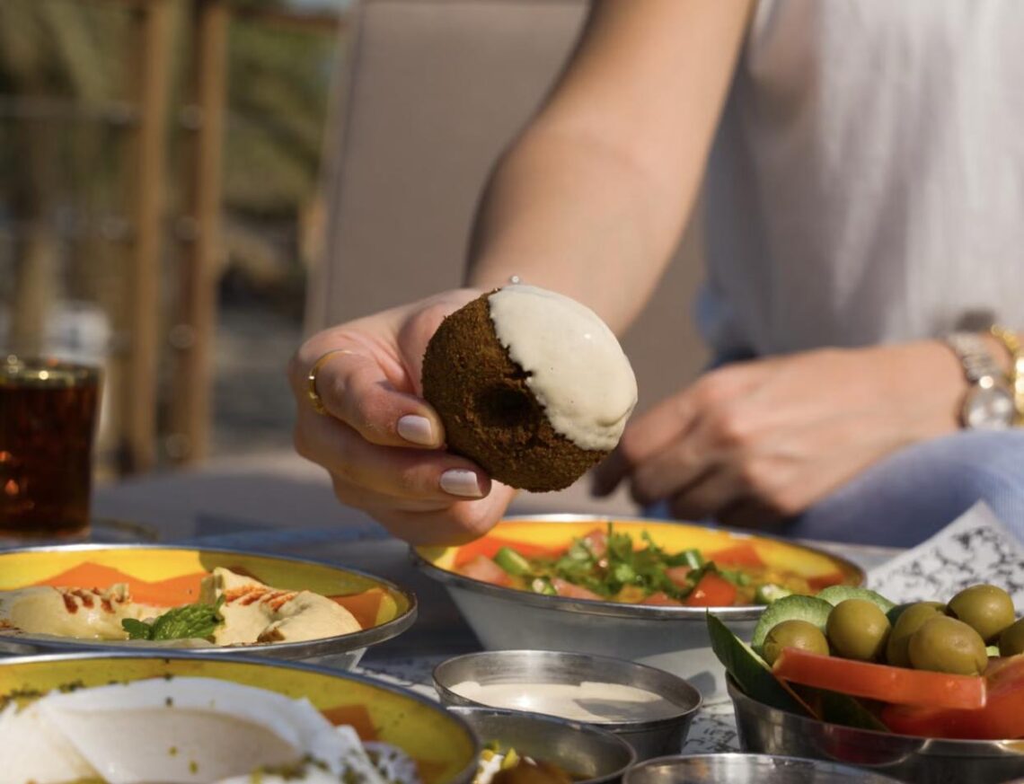 A hand holding a bitten falafel ball alongside a table spread of hummus, olives, and Middle Eastern dishes.