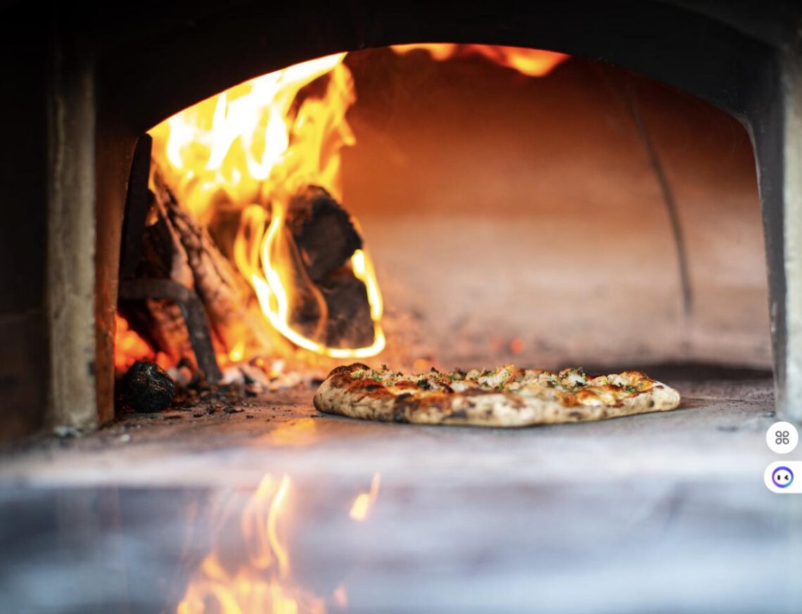 A Neapolitan-style pizza baking on the stone floor of a wood-fired oven with flames visible beside the charred logs.