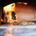 A Neapolitan-style pizza baking on the stone floor of a wood-fired oven with flames visible beside the charred logs.