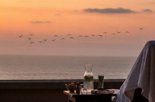 A flat lay of travel wellness items on a sandy beach background. The items include a passport, copper water bottle, CBD oil, sleep mask, and a phone with a yoga app. The text reads: "The 'How To' Guide: Weave Wellness Into Your Travels" and "The Ritz-Carlton Spa, Laguna Niguel."