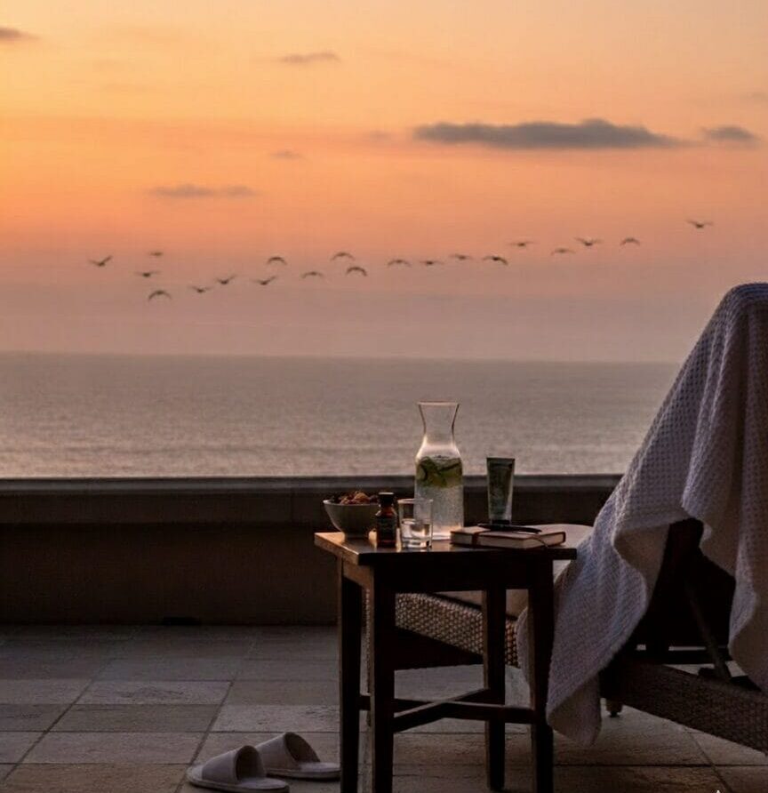 A flat lay of travel wellness items on a sandy beach background. The items include a passport, copper water bottle, CBD oil, sleep mask, and a phone with a yoga app. The text reads: "The 'How To' Guide: Weave Wellness Into Your Travels" and "The Ritz-Carlton Spa, Laguna Niguel."