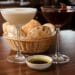 Two unique cocktails, one creamy white in a coupe glass and one dark burgundy in a wine glass, sit on a dark wooden table. In front of them is a woven basket filled with warm, puffy pita bread. The background is the soft, ambient lighting of the upscale Zaytinya restaurant