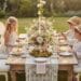 Three young children sit at a beautifully decorated wooden table for a whimsical, enchanted forest themed party in a sunlit garden. The table features a large floral centerpiece, tiered stands with cookies and macarons, and elegant place settings. A girl wears fairy wings.