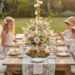 Three young children sit at a beautifully decorated wooden table for a whimsical, enchanted forest themed party in a sunlit garden. The table features a large floral centerpiece, tiered stands with cookies and macarons, and elegant place settings. A girl wears fairy wings.