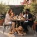 A man and a woman smile at each other while dining al fresco on a beautiful restaurant patio, while their golden doodle dog lies peacefully on a mat at their feet.