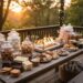 A beautifully styled outdoor gourmet S'mores dessert bar set up on a wooden deck at sunset. Glass jars hold various marshmallows and cookies, alongside different types of chocolate bars and a central tabletop fire pit. String lights glow in the background.