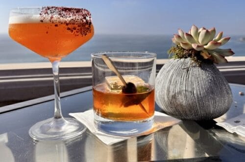 Two cocktails, one orange with a red rim in a coupe glass and one amber in an old-fashioned glass, sit on a glass table next to a succulent plant with the Pacific Ocean and a clear blue sky in the background, labeled "180blu - The Ritz Carlton, Laguna Niguel."