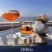 Two cocktails, one orange with a red rim in a coupe glass and one amber in an old-fashioned glass, sit on a glass table next to a succulent plant with the Pacific Ocean and a clear blue sky in the background, labeled "180blu - The Ritz Carlton, Laguna Niguel."