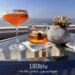 Two cocktails, one orange with a red rim in a coupe glass and one amber in an old-fashioned glass, sit on a glass table next to a succulent plant with the Pacific Ocean and a clear blue sky in the background, labeled "180blu - The Ritz Carlton, Laguna Niguel."