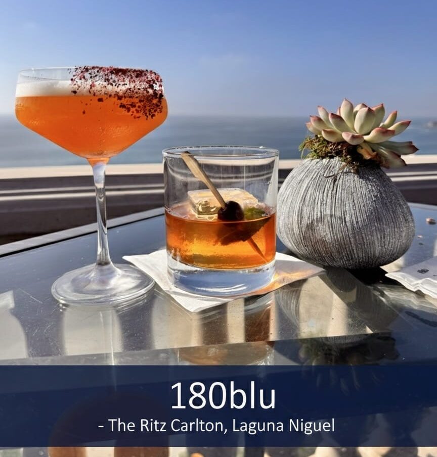 Two cocktails, one orange with a red rim in a coupe glass and one amber in an old-fashioned glass, sit on a glass table next to a succulent plant with the Pacific Ocean and a clear blue sky in the background, labeled "180blu - The Ritz Carlton, Laguna Niguel."