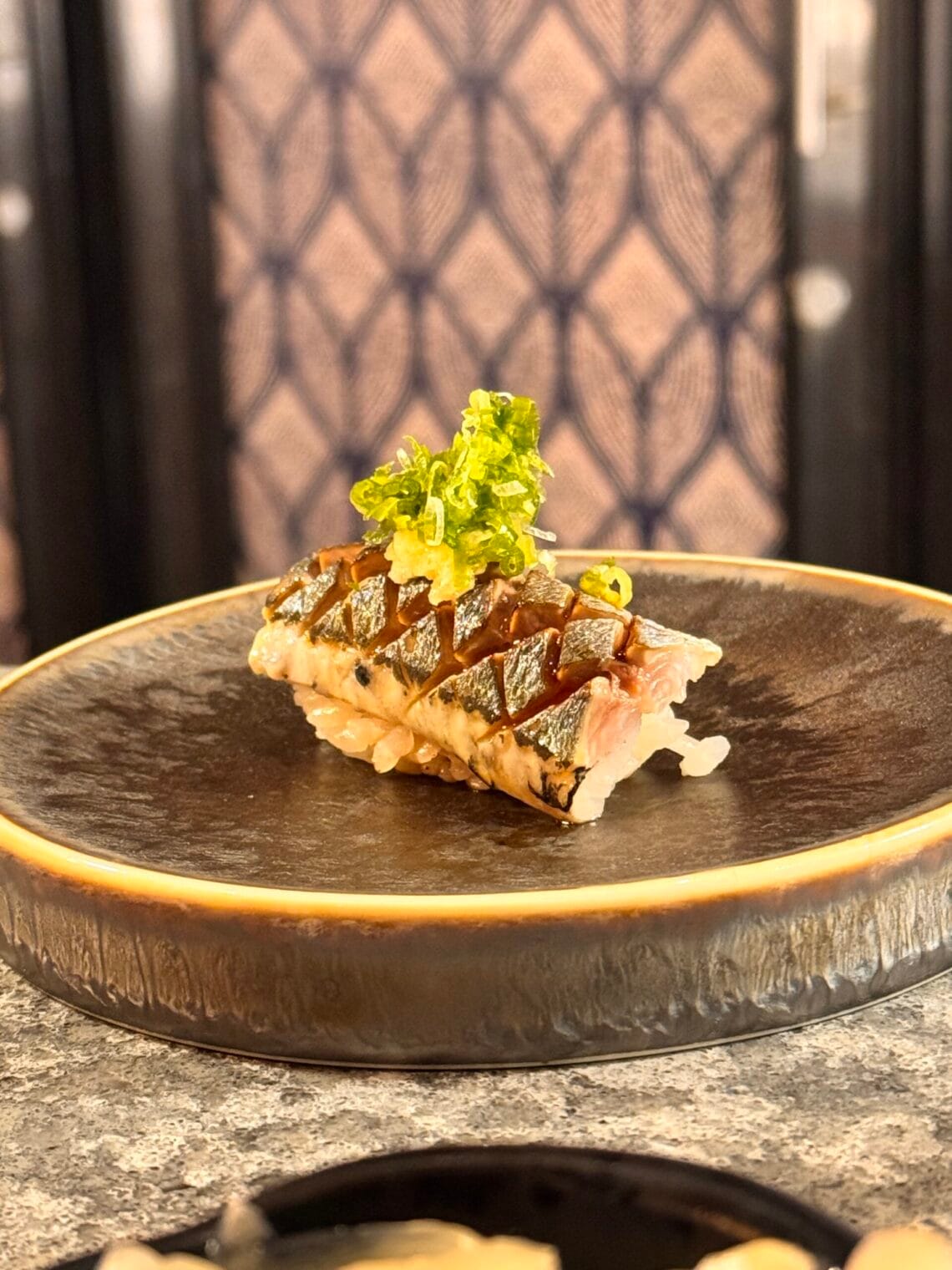 Close-up of a beautifully plated piece of seared nigiri, topped with vibrant green onions, on a dark ceramic dish.