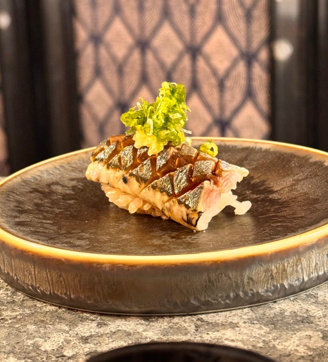 Close-up of a beautifully plated piece of seared nigiri, topped with vibrant green onions, on a dark ceramic dish.