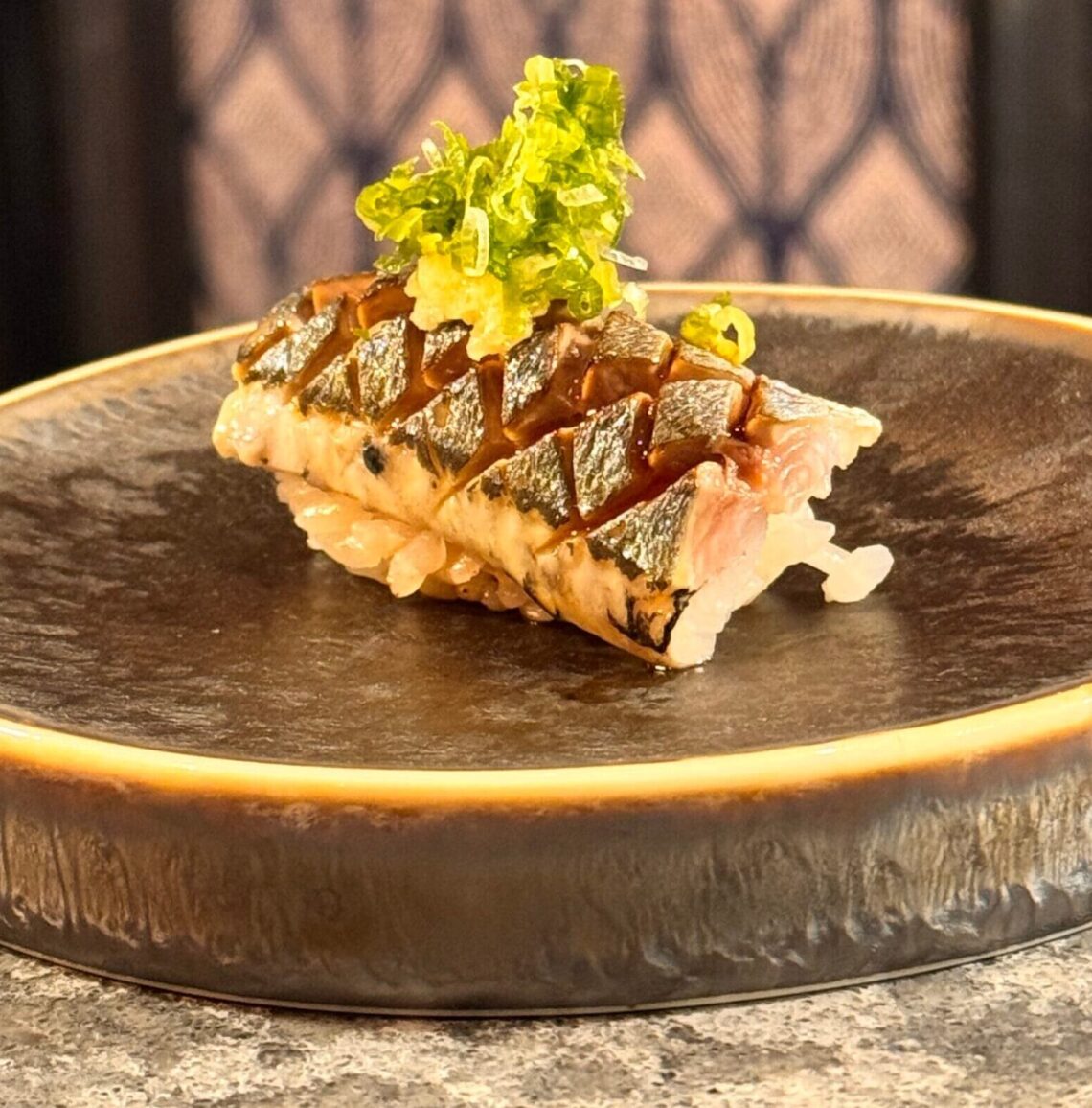 Close-up of a beautifully plated piece of seared nigiri, topped with vibrant green onions, on a dark ceramic dish.