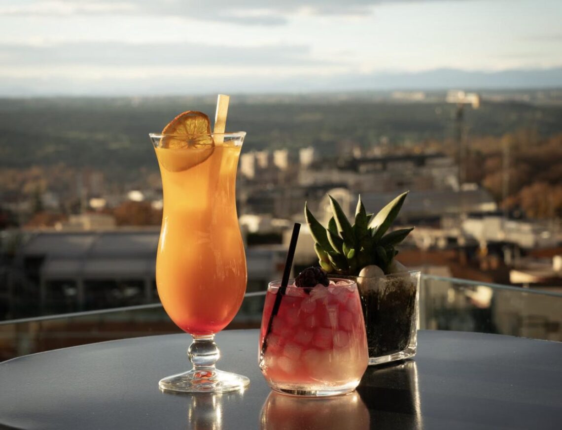 Two colorful cocktails on a rooftop bar table overlooking the city at sunset.