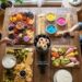 A vibrant overhead view of a rustic wooden table laden with five themed food boards for a "Bring a Board" party, including savory, sweet, and deconstructed dessert options, with friends toasting with wine glasses.
