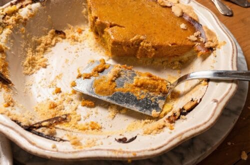 A half-eaten pumpkin pie in a scalloped ceramic dish resting on a marble trivet. A silver pie server sits in the dish with crumbly crust, while forks and a bowl of whipped cream sit in the background on a wooden table.