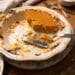 A half-eaten pumpkin pie in a scalloped ceramic dish resting on a marble trivet. A silver pie server sits in the dish with crumbly crust, while forks and a bowl of whipped cream sit in the background on a wooden table.
