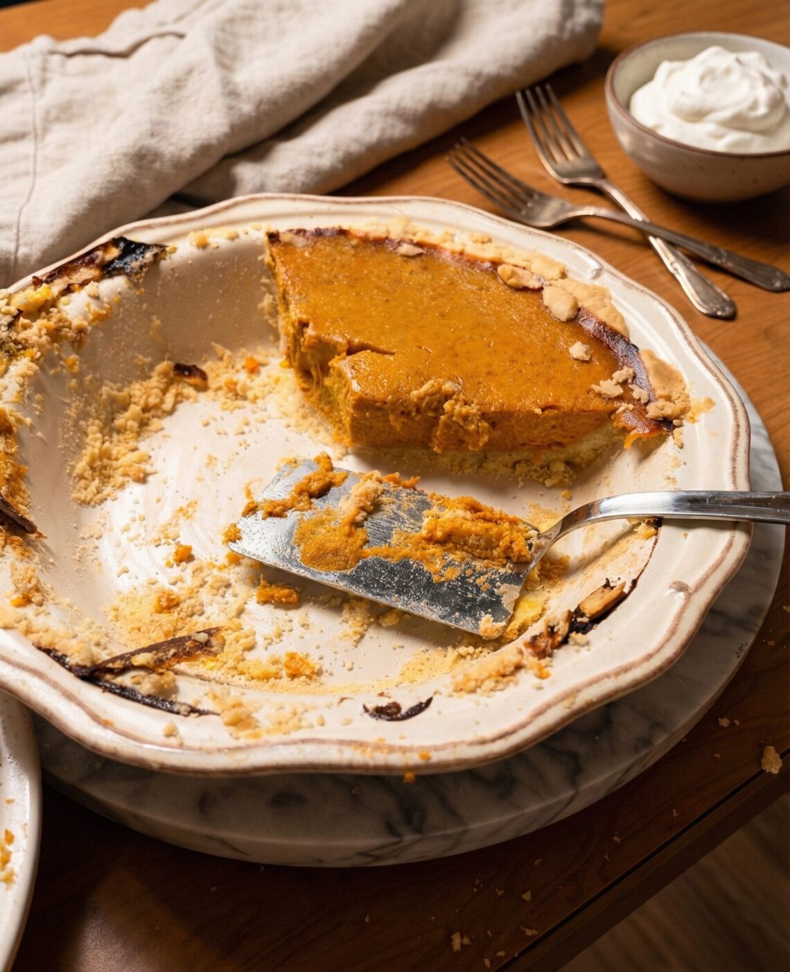 A half-eaten pumpkin pie in a scalloped ceramic dish resting on a marble trivet. A silver pie server sits in the dish with crumbly crust, while forks and a bowl of whipped cream sit in the background on a wooden table.