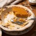 A half-eaten pumpkin pie in a scalloped ceramic dish resting on a marble trivet. A silver pie server sits in the dish with crumbly crust, while forks and a bowl of whipped cream sit in the background on a wooden table.