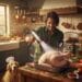 A smiling man in a green shirt uses a silver high-tech hairdryer on a fully roasted, golden-brown Thanksgiving turkey. As he aims the hairdryer, small white feathers float around the bird. Beside him, a golden retriever in a chef's hat looks on.