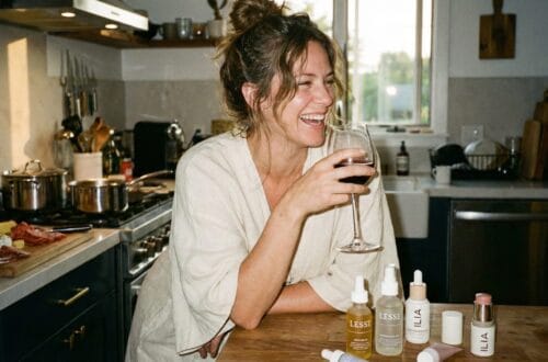 Ginger Graham laughing in a warm kitchen holding a glass of red wine, leaning on a wooden counter with Lesse Skincare bottles (Ritual Serum, Regeneration Mist) and ILIA Beauty makeup products (Super Serum Skin Tint, Multi Stick) displayed in front of her. A charcuterie board is visible in the background.