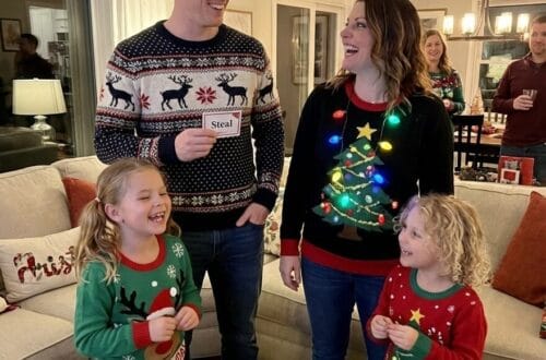 A family of four, wearing festive Christmas sweaters, laughs together around a coffee table piled with wrapped gifts. The father holds a card that says "Steal," and the mother's sweater features a light-up Christmas tree.