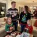 A family of four, wearing festive Christmas sweaters, laughs together around a coffee table piled with wrapped gifts. The father holds a card that says "Steal," and the mother's sweater features a light-up Christmas tree.