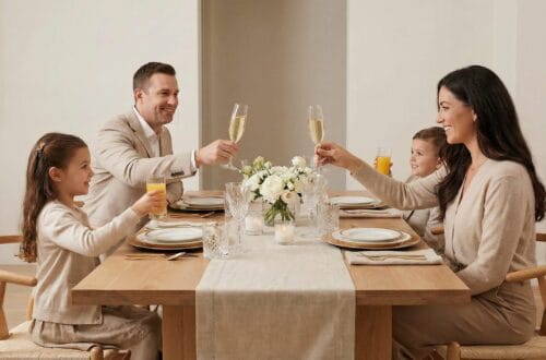 A family of four with two parents and two young children sit at a light wood dining table in a modern, neutral-toned dining room. They are all raising glasses, including champagne flutes and juice glasses, for a toast. A white floral arrangement and candles are on the table.