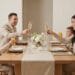 A family of four with two parents and two young children sit at a light wood dining table in a modern, neutral-toned dining room. They are all raising glasses, including champagne flutes and juice glasses, for a toast. A white floral arrangement and candles are on the table.