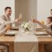 A family of four with two parents and two young children sit at a light wood dining table in a modern, neutral-toned dining room. They are all raising glasses, including champagne flutes and juice glasses, for a toast. A white floral arrangement and candles are on the table.