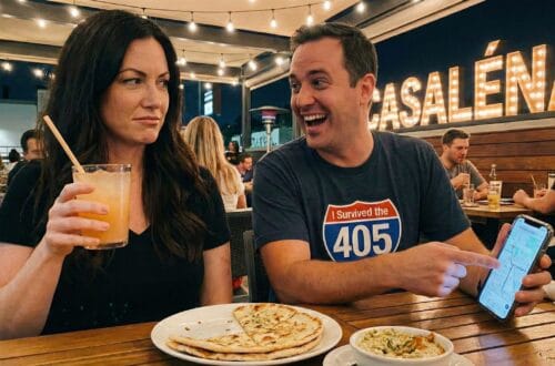 Ginger Graham (brunette woman) and Brad Graham (man) dining at Casaléna restaurant in Woodland Hills. Ginger looks skeptical holding a cocktail while Brad smiles broadly, holding a smartphone showing a map. Outdoor patio dining setting with string lights.