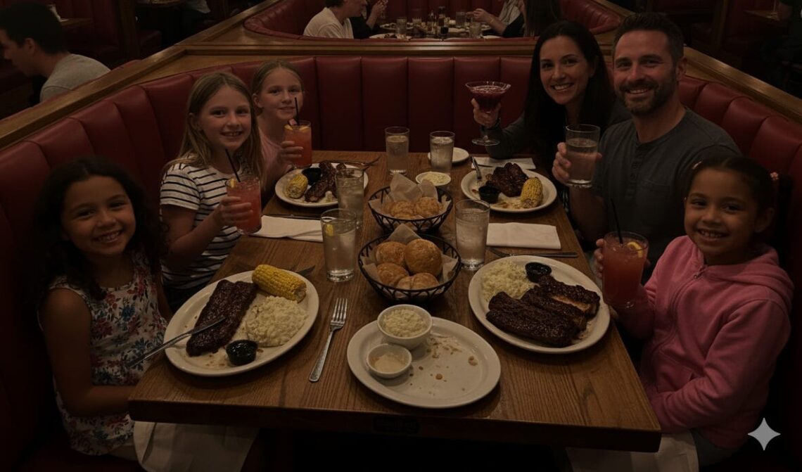 A family and their friends, two adults and four girls, happily eating ribs in a cozy, dimly lit red booth at Wood Ranch BBQ, with the mom holding a martini and kids drinking strawberry lemonade.