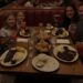 A family and their friends, two adults and four girls, happily eating ribs in a cozy, dimly lit red booth at Wood Ranch BBQ, with the mom holding a martini and kids drinking strawberry lemonade.
