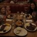 A family and their friends, two adults and four girls, happily eating ribs in a cozy, dimly lit red booth at Wood Ranch BBQ, with the mom holding a martini and kids drinking strawberry lemonade.