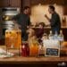A "Spike Station" for Thanksgiving cocktails on a table, including a large dispenser of apple cider, two pitchers, and bottles of bourbon, gin, and vodka. In the blurry background, a chaotic family scene unfolds in a kitchen.