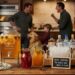 A "Spike Station" for Thanksgiving cocktails on a table, including a large dispenser of apple cider, two pitchers, and bottles of bourbon, gin, and vodka. In the blurry background, a chaotic family scene unfolds in a kitchen.