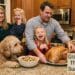 A candid Thanksgiving photo in a kitchen showing the Graham family – Ginger with a glass of wine, Brad holding a cooked turkey, daughters Madeline and Charlotte laughing, and their goldendoodle Barnaby eyeing a bowl of stuffing. A text overlay in the bottom right reads "Graham Family Thanksgiving Survival Guide."